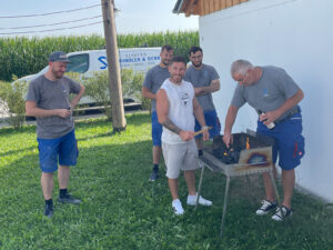 Eine Gruppe von fünf Männern, einige davon in blauen Shorts und grauen Hemden, steht draußen um einen Grill herum. Ein Mann hält eine Zange und kümmert sich um den Grill, während ein anderer Kohle nachlegt. Sie stehen auf einer Wiese, im Hintergrund sind ein Gebäude und ein Maisfeld zu sehen.